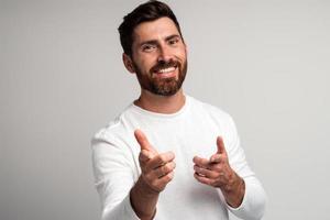 Hey you. Portrait of happy bearded man in white shirt smiling and pointing to the camera against light gray background. Studio shot photo