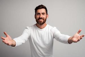 Hombre generoso de corazón abierto con barba compartiendo la apertura de las manos mirando a la cámara con una sonrisa amable, saludo y regocijo, feliz, feliz de verte. Foto de estudio de interior aislado sobre fondo blanco.