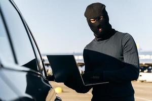 Masked thief in a balaclava stealing laptop from car and holding it opened while standing near the car. Criminal concept. Stock photo