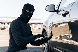 Waist up portrait of the car thief with laptop hacking alarm system while sitting near the car at the daytime. Stock photo