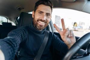 Making selfie. Handsome bearded man holding camera and gesturing while sitting in the car at the driver seat and taking self portrait with pleasure smile