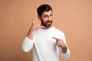 Llámame. Retrato de hombre guapo alegre de pie con gesto de la mano del teléfono y sonriendo a la cámara, coqueteando ofreciendo contactar por teléfono. Foto de estudio aislado sobre fondo beige