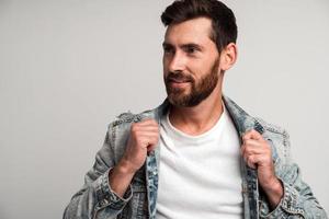 Retrato de hombre guapo con barba en estilo casual mirando a otro lado y sonriendo mientras se endereza la ropa y posa para la cámara. Foto de estudio con fondo blanco