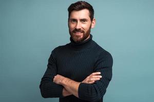 Portrait of happy satisfied handsome young man standing with crossed arms and looking at camera with toothy smile. Indoor studio shot on blue background copy space photo