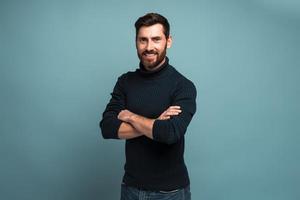 Portrait of happy successful handsome bearded young man standing with crossed arms and looking at camera with smile. Indoor studio shot, isolated on blue background photo