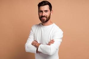 Retrato de guapo feliz joven barbudo de pie con los brazos cruzados y mirando a la cámara con una gran sonrisa. Foto de estudio de interior aislado sobre fondo beige