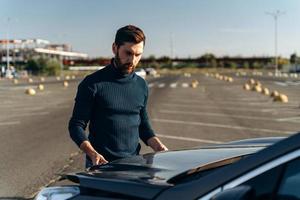 Upset bearded man checking his car engine after breaking down on the road. Serious male is standing in front of the hood. Transportation concept photo