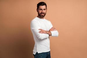 Retrato de un joven barbudo satisfecho guapo con camisa blanca de pie con los brazos cruzados y mirando a la cámara con una sonrisa. Foto de estudio de interior aislado sobre fondo beige