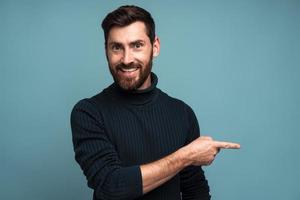 Advertise here. Positive bearded man pointing finger away paying your attention at empty space for advertisement, looking at camera with toothy smile. Indoor studio shot isolated on blue background photo