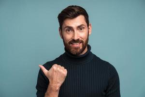 mira aquí. Retrato de hombre moreno amable positivo con barba de pie y apuntando a la izquierda, espacio vacío para texto, publicidad. Foto de estudio de interior aislado sobre fondo azul.