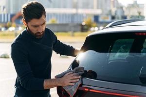 Waist up portrait view of a satisfied concentrated man cleaning the back of his new car while spending time at the street during the sunny summer day. Stock photo