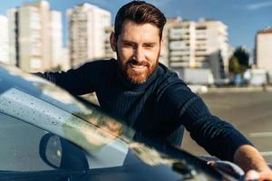 Smiling man cleaning car and drying vehicle with microfiber cloth. Hand wipe down paint surface of shiny car after polishing. Car detailing and car wash concept photo