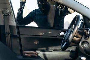 vista desde el auto al hombre vestido de negro con un pasamontañas en la cabeza preparándose para romper con una palanca el vidrio del auto antes del robo. ladrón de autos, concepto de robo de autos foto