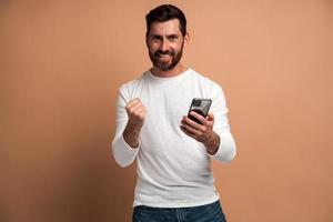 Hombre feliz satisfecho con barba sosteniendo teléfono inteligente y sonriendo haciendo gesto de sí, celebrando la lotería en línea o la victoria del sorteo. Foto de estudio de interior aislado sobre fondo beige