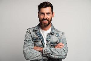 Hombre barbudo adulto joven con chaqueta casual posando con expresión facial segura y brazos cruzados, mirando sonriendo a la cámara. Foto de estudio de interior aislado sobre fondo blanco.