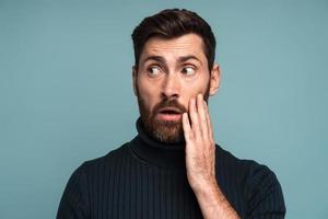 Excited shocked man holding hands on face and looking away with big eyes and open mouth, shocked bemused with news, win at lottery. Indoor studio shot isolated on blue background photo