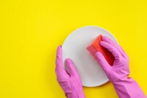 Closeup hands wearing in latex gloves holding a kitchen sponge and plate.  Top view on yellow  background washing dishes photo