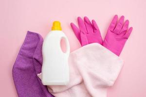 Botella de detergente con tapón amarillo, guantes de látex y trapo sobre un fondo rosa foto