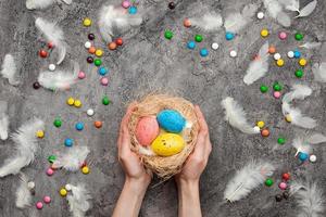 Fondo de Pascua, manos femeninas sosteniendo un nido con huevos multicolores. dulces y plumas esparcidas
