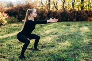 Beautiful girl in black sportswear performs squats in the park, side view. photo