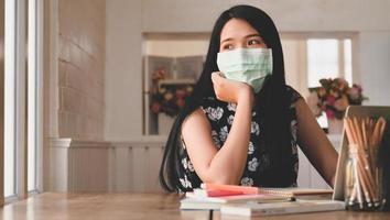 Tired woman wearing a medical mask looks out the window, with laptop and stationery on the table. photo