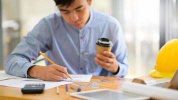 joven diseñador con una taza de café para llevar en la mano está usando un lápiz para dibujar el diseño de una casa. foto