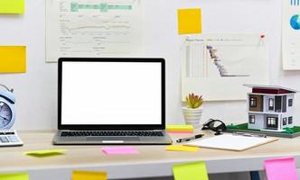 Blank screen laptop mockup with clipboard, model house with office supplies on the table and graph data on the wall. photo