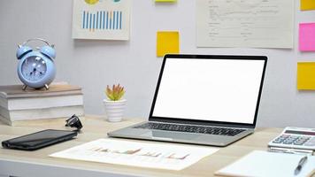 Mockup Blank screen laptop with graph, calculator, smartphone on the desk, clock on the book, chart on back wall, Office desk. photo