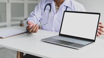 un hombre vestido con una bata médica, sostiene un bolígrafo con una pantalla de computadora portátil en blanco al frente.