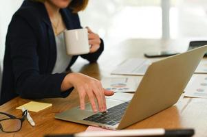 una mujer en traje presiona su mano sobre el teclado de una computadora portátil y sostiene el café con la otra mano.
