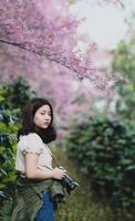 Adolescente asiática con una cámara se encuentra mirándola debajo de un árbol de cerezos en flor. foto