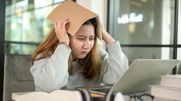 Asian teenage girl holding a book on her head stressed looking at laptop during online lessons from laptop. photo