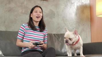 A casual Asian young woman is playing a video game console with fun and excitement controlled by a joystick on a sofa in the home's living room. The pet is a dog, French Bulldog looking close.