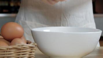 images de vue de face en gros plan, une cuisinière en tablier blanc casse un œuf dans une tasse pour préparer un repas sur une table en bois dans la cuisine de la maison. manger des jaunes d'œufs est un petit-déjeuner sain. video