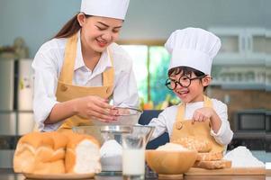 Cute little Asian boy and beautiful mother sifting dough flour with sifter sieve colander in home kitchen on table for prepare to baking bakery and cake. Thai kids playing with flour as chef funny photo