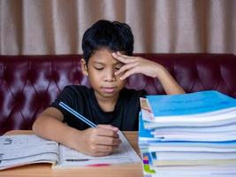 boy doing homework and reading on a wooden table with a pile of books beside The background is a red sofa and cream curtains. photo