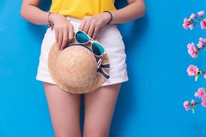 Closeup of Beauty woman posing with sunglasses and straw hat in front of blue wall background. Summer and vintage concept. Happiness lifestyle and people portrait theme. Cute pastel tone. Lower body photo
