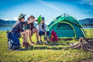 Group of young Asian friends enjoy picnic and party at lake with camping backpack and chair. Young people toasting and cheering bottles of beer. People and lifestyles concept. Outdoor background theme photo