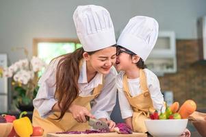 Happy cute little boy with eyeglasses kissing beautiful Asian woman mother while prepare to cooking in kitchen at home. People lifestyles and Family. Homemade food and ingredients concept