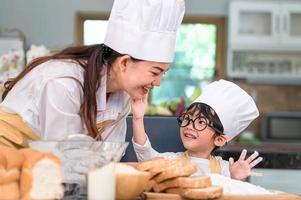 Cute little Asian boy painting beautiful woman face with dough flour. Chef team playing and baking bakery in home kitchen funny. Homemade food and bread. Education teamwork and learning concept photo