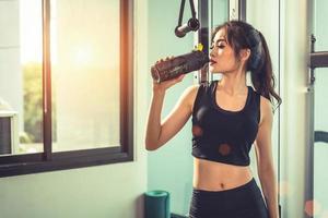 Asian young woman drinking protein shake or water after exercises at fitness gym. Strength training and muscular. Beauty and Healthy concept. Relax concept. Sport equipment and Club center theme photo