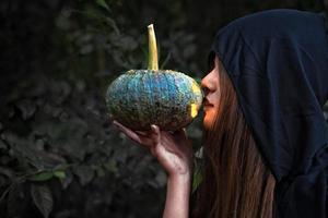 maga besando linterna de calabaza. traje femenino con ropa negra y capucha sobre fondo de bosque misterioso. día de halloween y concepto de misterio. fantasía de tema mágico. concepto de vida después de la muerte y muerte foto