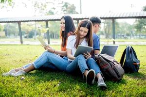Group of Asian college student using tablet and laptop on grass field at outdoors. Technology and Education learning concept. Future Technology and Modern entertainment concept. Edutainment theme