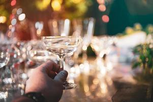 Primer plano mano sujetando copas de vino de martini en la barra de mostrador al aire libre en el jardín por la noche para la fiesta de celebración foto