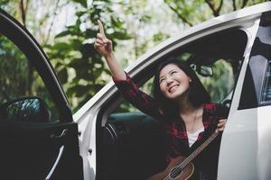 Beauty Asian woman pointing and having fun at outdoors summer with Ukulele in white car. Traveling of photographer concept. Hipster style and Solo woman theme. Lifestyle and Happiness life theme photo