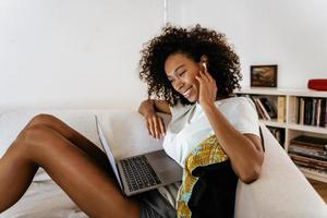 Black young woman with earphones using laptop while resting on sofa photo
