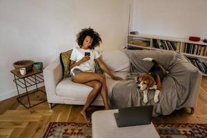 Black young woman on home sofa using mobile phone while resting with her dog  photo