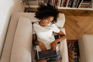 Black young woman in earphones using laptop while resting on sofa