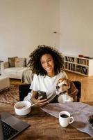 Mujer negra con teléfono celular y abrazando a su perro mientras desayuna foto