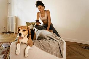 Black young woman using cellphone while sitting with her dog on sofa photo